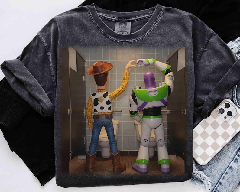 May include: A charcoal gray mineral-washed t-shirt featuring a graphic print of Woody and Buzz Lightyear standing in bathroom stalls, forming a heart shape with their hands. The characters are shown from behind, wearing their signature outfits.