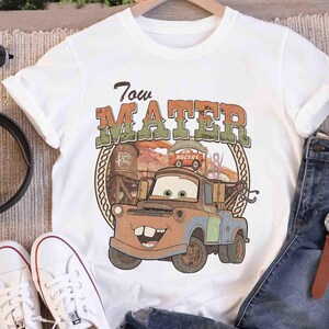 K&ouml;nnte beinhalten: Ein wei&szlig;es Kurzarm-T-Shirt mit einem Grafikdruck der Figur Tow Mater aus dem Film Cars. Das Design enth&auml;lt den Text Tow Mater in einer abgenutzten Schriftart, einen W&uuml;stenhintergrund und eine kreisf&ouml;rmige Umrandung im Seil-Stil.