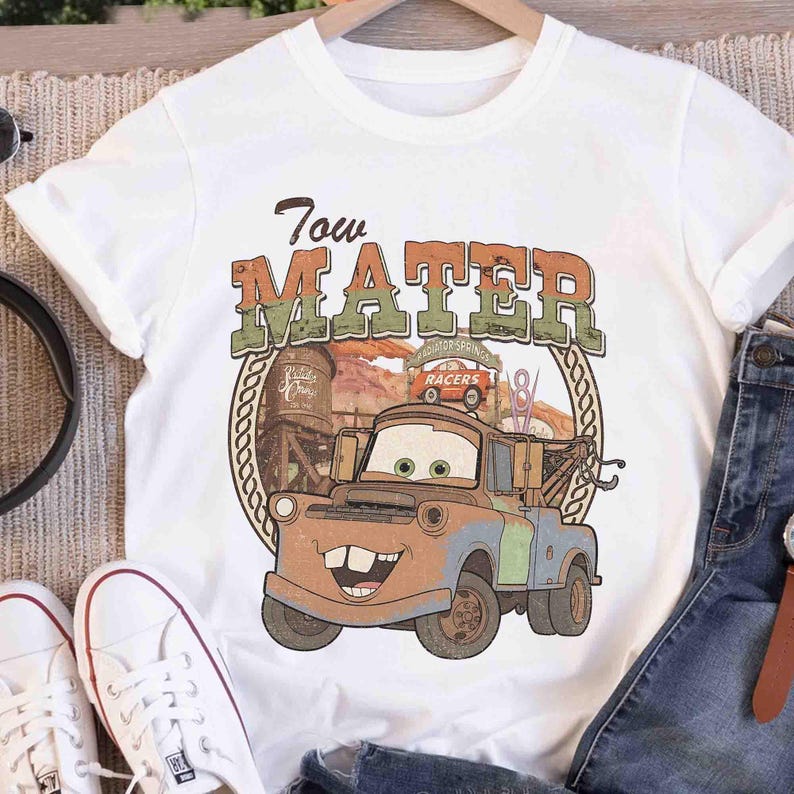 K&ouml;nnte beinhalten: Ein wei&szlig;es Kurzarm-T-Shirt mit einem Grafikdruck der Figur Tow Mater aus dem Film Cars. Das Design enth&auml;lt den Text Tow Mater in einer abgenutzten Schriftart, einen W&uuml;stenhintergrund und eine kreisf&ouml;rmige Umrandung im Seil-Stil.