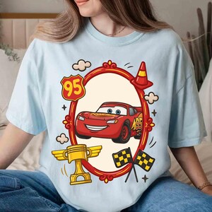 May include: Light blue t-shirt featuring a cartoon image of a red race car with the number 95. The design includes a gold trophy, checkered flags, and a traffic cone. The car is inside a red oval frame with decorative elements.