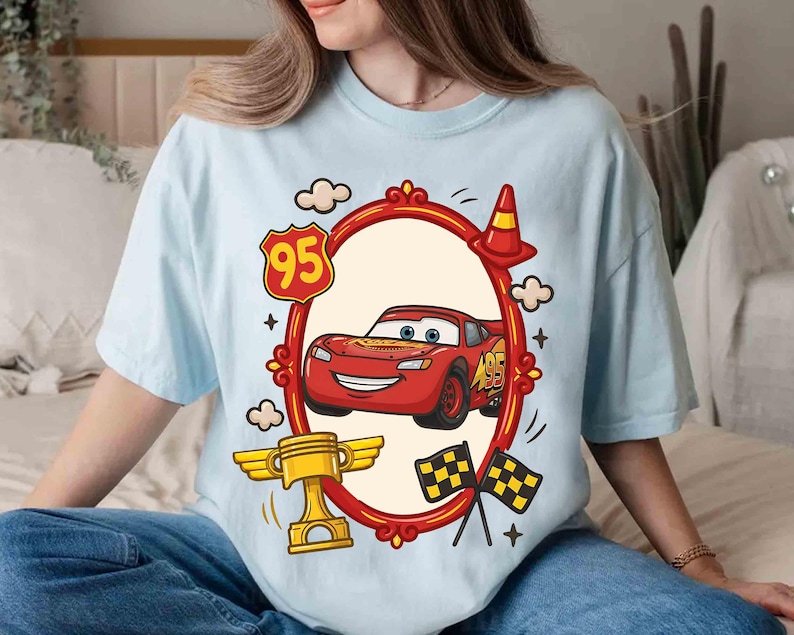 May include: Light blue t-shirt featuring a cartoon image of a red race car with the number 95. The design includes a gold trophy, checkered flags, and a traffic cone. The car is inside a red oval frame with decorative elements.