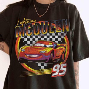 May include: Black t-shirt featuring Lightning McQueen from the Cars film. The design includes a red race car with the number 95, set against a checkered flag background, and the text "Lightning McQueen".