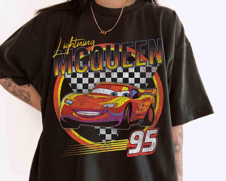 May include: Black t-shirt featuring Lightning McQueen from the Cars film. The design includes a red race car with the number 95, set against a checkered flag background, and the text "Lightning McQueen".