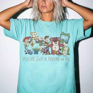 Op de afbeelding: Een lichtblauw T-shirt met een kleurrijke afbeelding van personages uit de animatiefilm "Toy Story". Het ontwerp bevat Woody, Buzz Lightyear en andere geliefde personages. De tekst "YOU'VE GOT A FRIEND IN ME" staat eronder.