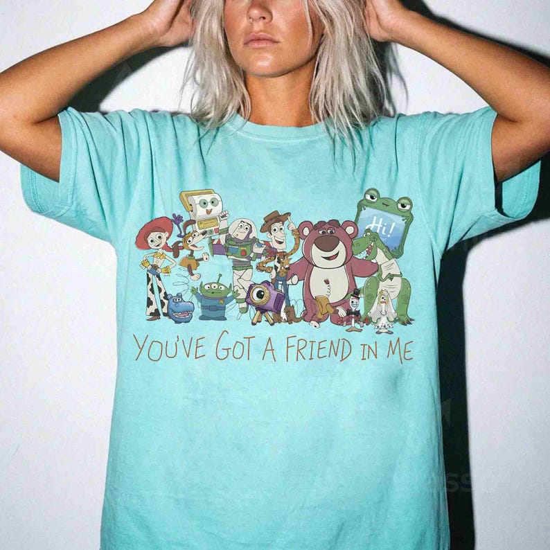 Op de afbeelding: Een lichtblauw T-shirt met een kleurrijke afbeelding van personages uit de animatiefilm "Toy Story". Het ontwerp bevat Woody, Buzz Lightyear en andere geliefde personages. De tekst "YOU'VE GOT A FRIEND IN ME" staat eronder.