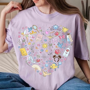 May include: A light purple t-shirt featuring a heart-shaped design filled with colorful illustrations of fairy tale elements. The design includes a fairy, a crown, a magic lamp, a rose, and other whimsical icons.