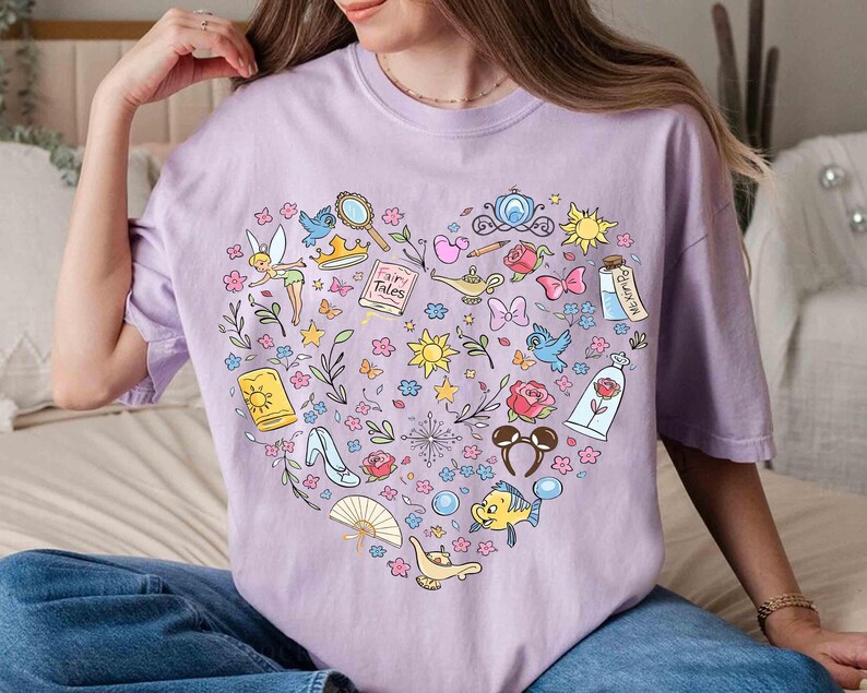 May include: A light purple t-shirt featuring a heart-shaped design filled with colorful illustrations of fairy tale elements. The design includes a fairy, a crown, a magic lamp, a rose, and other whimsical icons.