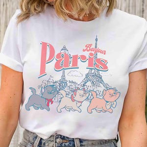 May include: White t-shirt featuring the word "Paris" in pink and blue, with "Bonjour" above. The design includes the Eiffel Tower and three cartoon cats. The cats are in gray, pink, and peach colors.