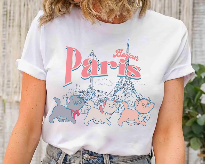 May include: White t-shirt featuring the word "Paris" in pink and blue, with "Bonjour" above. The design includes the Eiffel Tower and three cartoon cats. The cats are in gray, pink, and peach colors.