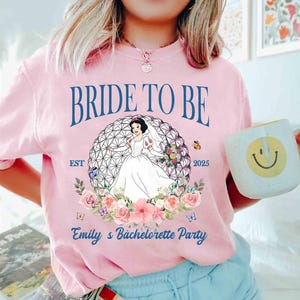 K&ouml;nnte beinhalten: Hellrosa T-Shirt mit dem Text "BRIDE TO BE" und "Emily's Bachelorette Party". Das Shirt zeigt eine Grafik einer Braut in einem wei&szlig;en Kleid in einem geometrischen Design, umgeben von Blumen und Schmetterlingen. Eine Tasse mit einem Smiley wird in der Hand gehalten.