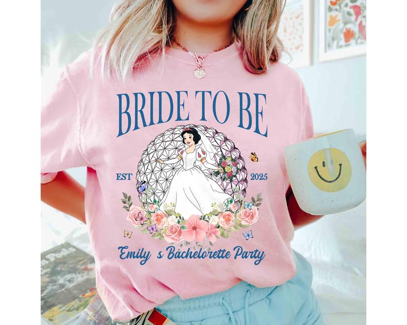 K&ouml;nnte beinhalten: Hellrosa T-Shirt mit dem Text "BRIDE TO BE" und "Emily's Bachelorette Party". Das Shirt zeigt eine Grafik einer Braut in einem wei&szlig;en Kleid in einem geometrischen Design, umgeben von Blumen und Schmetterlingen. Eine Tasse mit einem Smiley wird in der Hand gehalten.
