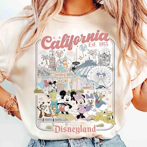 Puede incluir: Camiseta color crema con un gr&aacute;fico colorido de Disneyland en California. El dise&ntilde;o incluye lugares emblem&aacute;ticos, fuegos artificiales y personajes de Disney como Mickey Mouse, Minnie Mouse y Donald Duck. El texto dice "California Est. 1955" y "Disneyland".
