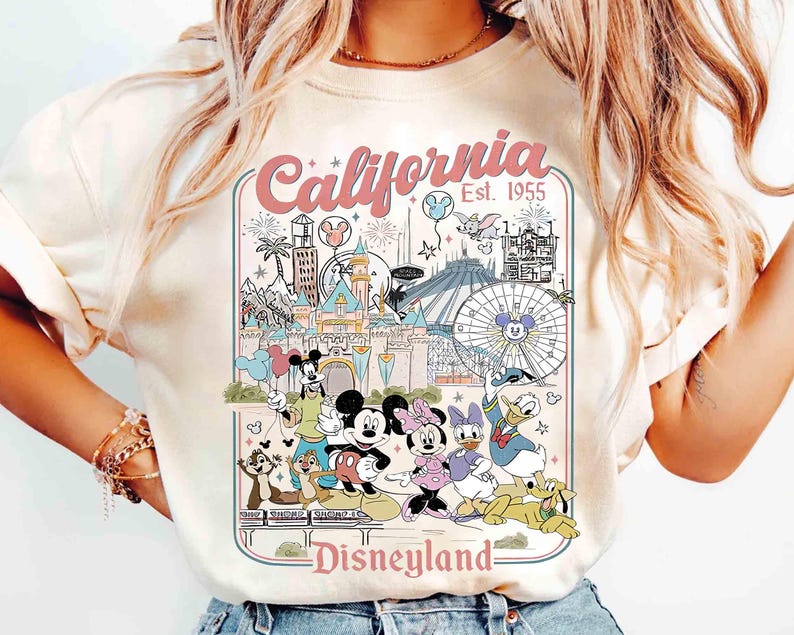 Puede incluir: Camiseta color crema con un gr&aacute;fico colorido de Disneyland en California. El dise&ntilde;o incluye lugares emblem&aacute;ticos, fuegos artificiales y personajes de Disney como Mickey Mouse, Minnie Mouse y Donald Duck. El texto dice "California Est. 1955" y "Disneyland".