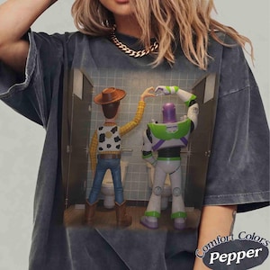 May include: A charcoal gray t-shirt featuring a graphic print of two toy characters standing in bathroom stalls. The character on the left wears a cowboy hat, yellow shirt, and blue jeans. The character on the right is a white, green, and purple space figure.