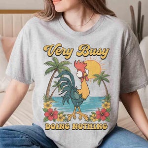 以下が含まれることがあります： 漫画のニワトリのデザインと「Very Busy Doing Nothing」の文字が入ったライトグレーのTシャツ。 ニワトリは黄色、青、赤で、ヤシの木、花、太陽のある熱帯の背景が描かれています。 文字は黄色と黒です。