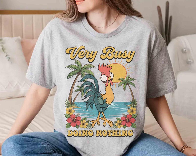 以下が含まれることがあります： 漫画のニワトリのデザインと「Very Busy Doing Nothing」の文字が入ったライトグレーのTシャツ。 ニワトリは黄色、青、赤で、ヤシの木、花、太陽のある熱帯の背景が描かれています。 文字は黄色と黒です。