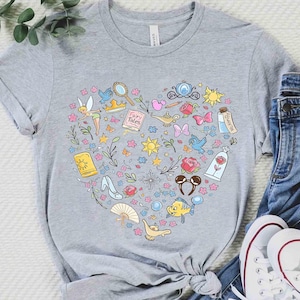 May include: A gray t-shirt featuring a heart-shaped design filled with colorful illustrations of fairy tale characters and objects. The design includes a fairy, a magic lamp, a glass slipper, and a rose. The shirt is paired with blue jeans and white sneakers.