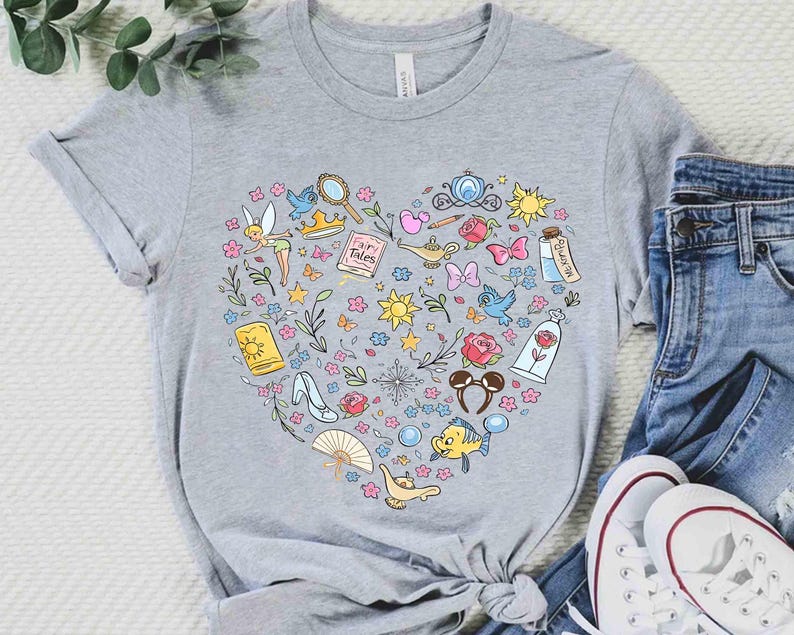 May include: A gray t-shirt featuring a heart-shaped design filled with colorful illustrations of fairy tale characters and objects. The design includes a fairy, a magic lamp, a glass slipper, and a rose. The shirt is paired with blue jeans and white sneakers.
