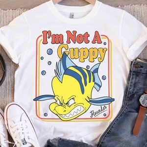 Op de afbeelding: Wit T-shirt met een cartoon illustratie van Bot uit De Kleine Zeemeermin. De tekst "I'm Not A Guppy" is in retro stijl weergegeven. De vis is geel met blauwe strepen en een boze uitdrukking.