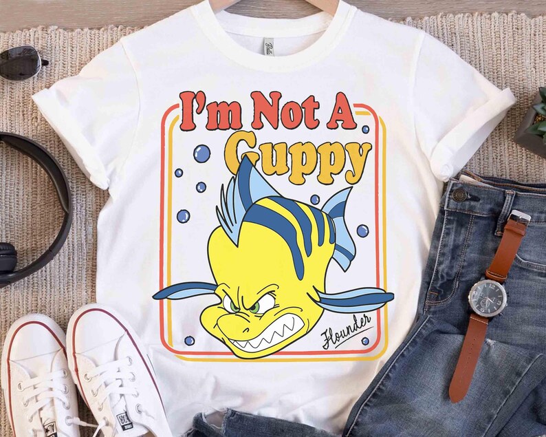 Op de afbeelding: Wit T-shirt met een cartoon illustratie van Bot uit De Kleine Zeemeermin. De tekst "I'm Not A Guppy" is in retro stijl weergegeven. De vis is geel met blauwe strepen en een boze uitdrukking.