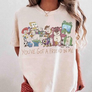 Op de afbeelding: Cr&egrave;mekleurig T-shirt met een kleurrijke afbeelding van Toy Story-personages. De tekst "You've Got a Friend in Me" staat in bruin schrift onder de personages. Het shirt heeft korte mouwen en is van zacht materiaal.