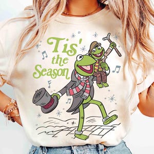 Puede incluir: Camiseta color crema con la Rana Gustavo en una escena invernal festiva. Gustavo lleva un sombrero de copa, abrigo y bufanda, con la frase "Tis' the Season" en verde. El diseño incluye notas musicales y copos de nieve.