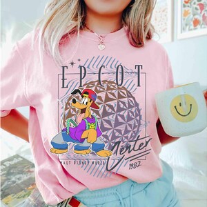 May include: A light pink t-shirt featuring a cartoon illustration of Pluto in a red cap, purple shirt, and blue jeans. The shirt has the text "EPCOT Center" and "1982" with a geometric sphere design, and is from Walt Disney World.