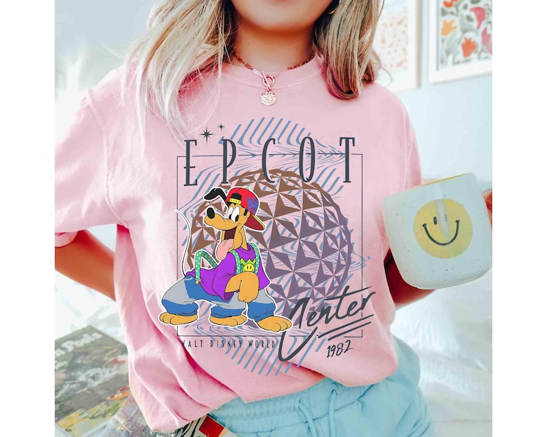 May include: A light pink t-shirt featuring a cartoon illustration of Pluto in a red cap, purple shirt, and blue jeans. The shirt has the text "EPCOT Center" and "1982" with a geometric sphere design, and is from Walt Disney World.