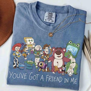 Op de afbeelding: Blauw Comfort Colors T-shirt met een afbeelding van Toy Story-personages en de tekst "You've Got a Friend in Me". Een gouden ketting met een hanger ligt op het T-shirt. Maat S-P-CH.