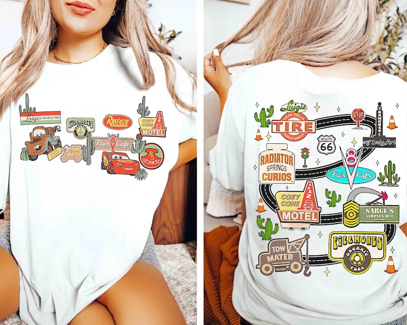 May include: White t-shirt featuring colorful graphics from the movie Cars. The front design includes characters and signs like Mater, Lightning McQueen, and the Cozy Cone Motel. The back design shows a road map with Route 66 and other iconic elements.