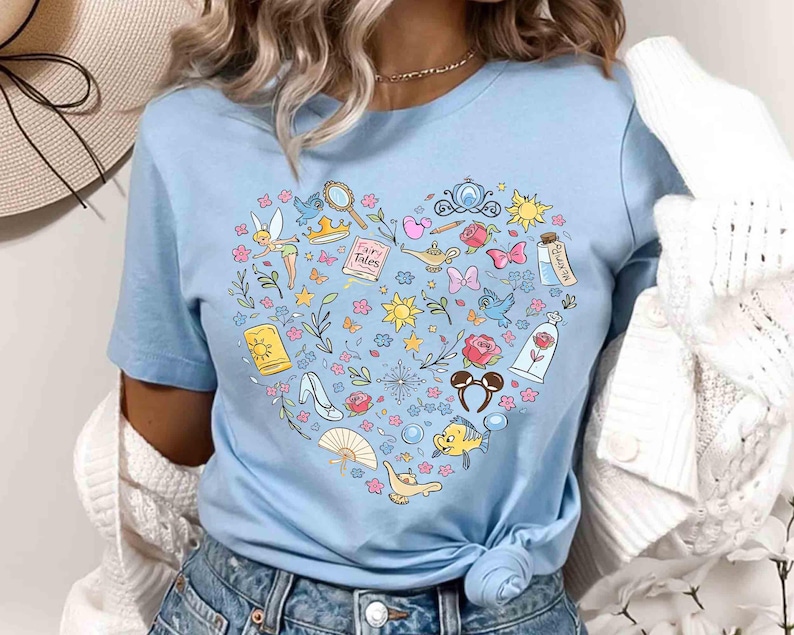 May include: Light blue t-shirt featuring a heart-shaped design filled with colorful illustrations of fairy tale elements. Includes a fairy, a magic lamp, a glass slipper, and the text "Fairy Tales".