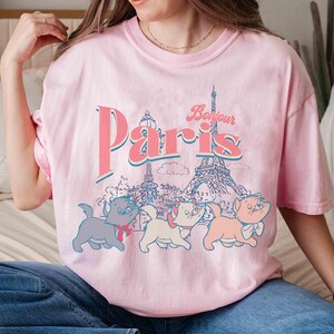 May include: Pink t-shirt with a graphic of the Eiffel Tower and the word "Paris" in a retro font. Below the text are three cartoon kittens. The shirt is a light pink color.