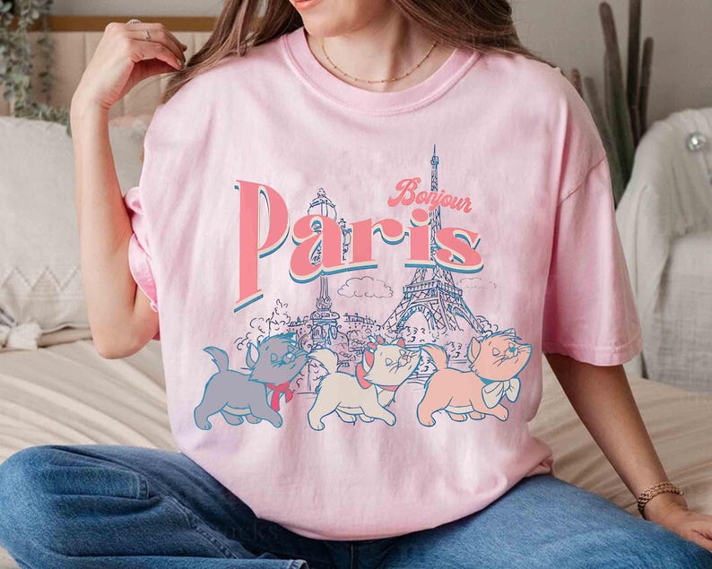 May include: Pink t-shirt with a graphic of the Eiffel Tower and the word "Paris" in a retro font. Below the text are three cartoon kittens. The shirt is a light pink color.
