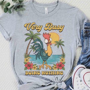 以下が含まれることがあります： 漫画の雄鶏が描かれたライトグレーのTシャツで、「Very Busy Doing Nothing」の文字が入っています。デザインには太陽、ヤシの木、花が含まれています。シャツは青いジーンズと白いスニーカーと組み合わせて表示されています。
