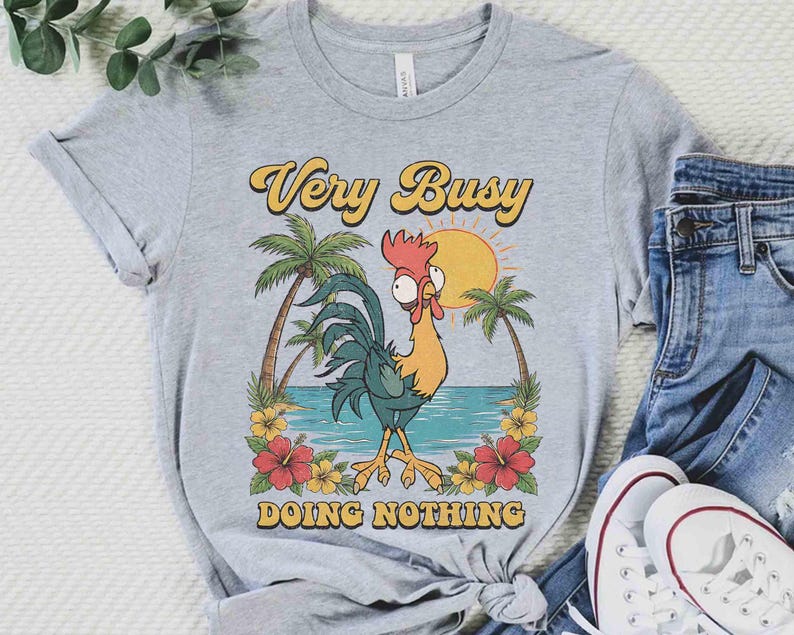 以下が含まれることがあります： 漫画の雄鶏が描かれたライトグレーのTシャツで、「Very Busy Doing Nothing」の文字が入っています。デザインには太陽、ヤシの木、花が含まれています。シャツは青いジーンズと白いスニーカーと組み合わせて表示されています。
