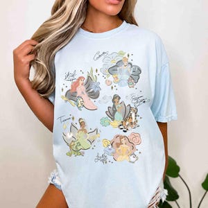 May include: A light blue t-shirt featuring a graphic print of six animated characters including Ariel, Cinderella, Jasmine, Tiana, and Belle. Each character is surrounded by thematic icons and their respective names written in a cursive script font.