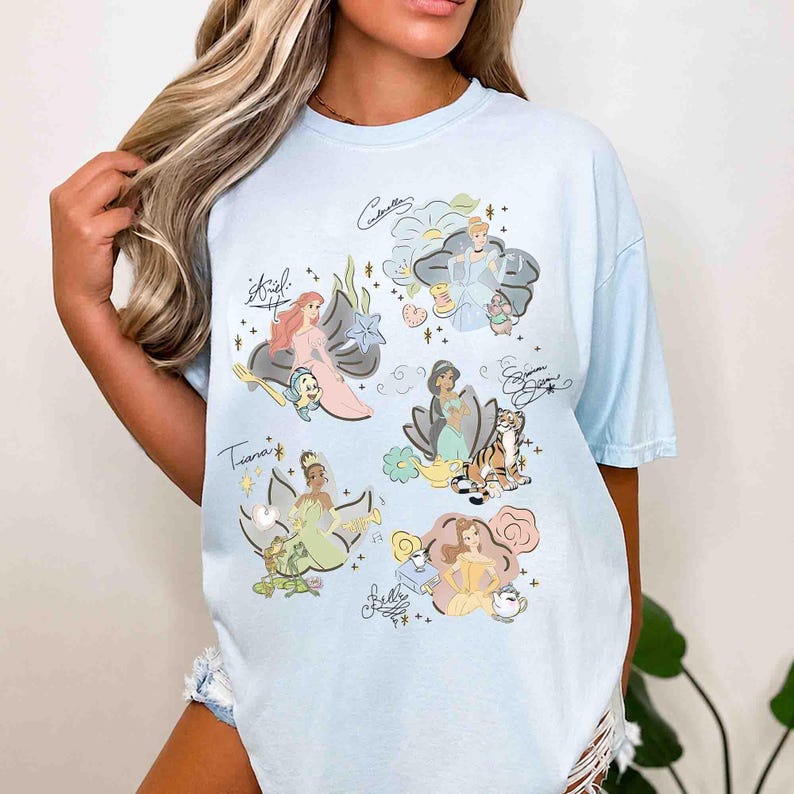 May include: A light blue t-shirt featuring a graphic print of six animated characters including Ariel, Cinderella, Jasmine, Tiana, and Belle. Each character is surrounded by thematic icons and their respective names written in a cursive script font.