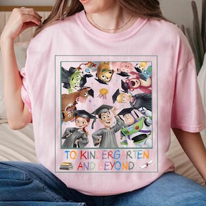 May include: A light pink t-shirt featuring a colorful graphic of characters from the movie Toy Story, wearing graduation caps. The text "TO KINDERGARTEN AND BEYOND" is printed below the graphic, with a stack of books and a graduation scroll.