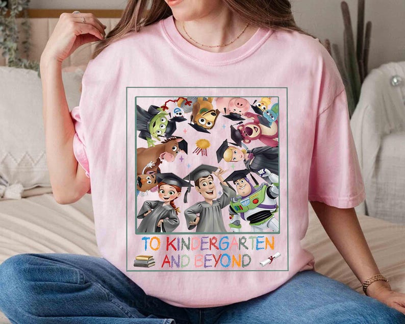 May include: A light pink t-shirt featuring a colorful graphic of characters from the movie Toy Story, wearing graduation caps. The text "TO KINDERGARTEN AND BEYOND" is printed below the graphic, with a stack of books and a graduation scroll.