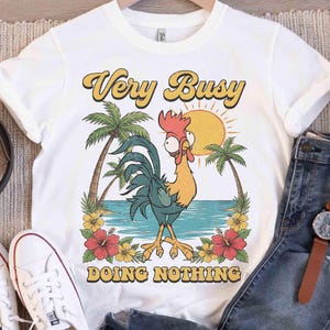 以下が含まれることがあります： 白いTシャツに漫画のニワトリのイラスト。デザインには、ヤシの木、太陽、花とともに、黄色と茶色で「Very Busy DOING NOTHING」というテキストが含まれています。このシャツは、青いジーンズと白いスニーカーと組み合わせています。
