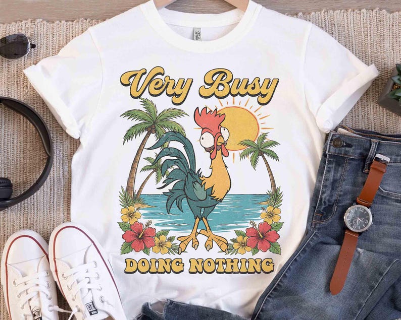 以下が含まれることがあります： 白いTシャツに漫画のニワトリのイラスト。デザインには、ヤシの木、太陽、花とともに、黄色と茶色で「Very Busy DOING NOTHING」というテキストが含まれています。このシャツは、青いジーンズと白いスニーカーと組み合わせています。