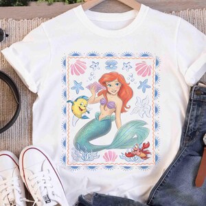 Op de afbeelding: Een wit t-shirt met korte mouwen en een centrale rechthoekige afbeelding van een zeemeermin met rood haar en een groene staart, een gele vis en een rode krab. Het ontwerp is omlijst door een blauwe rand met kleine zonnestralen.