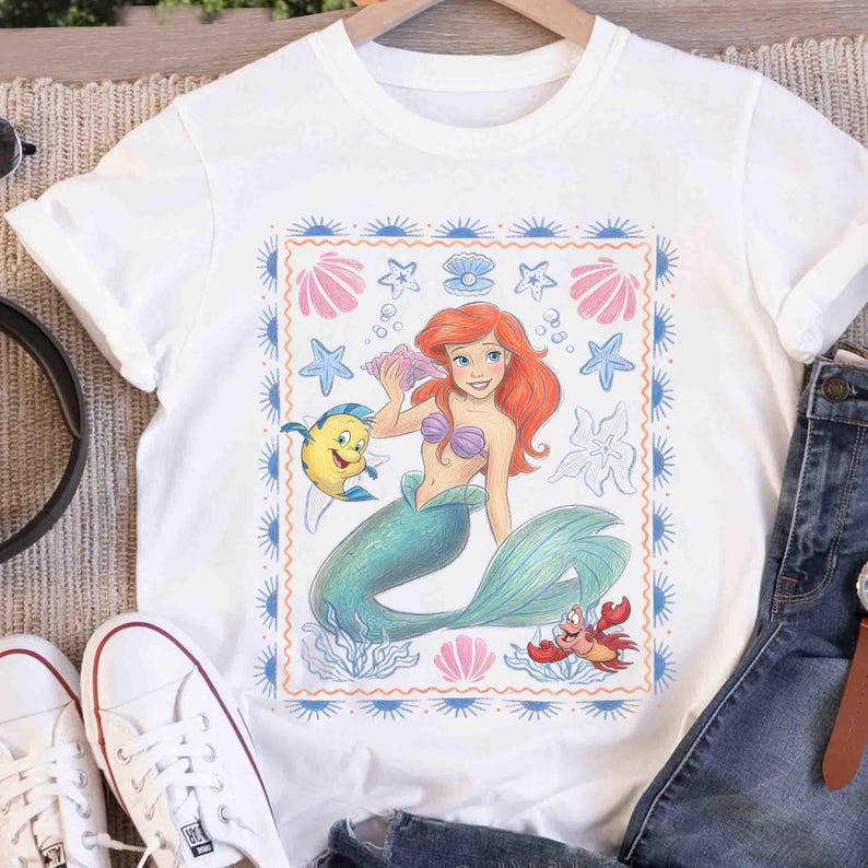 Op de afbeelding: Een wit t-shirt met korte mouwen en een centrale rechthoekige afbeelding van een zeemeermin met rood haar en een groene staart, een gele vis en een rode krab. Het ontwerp is omlijst door een blauwe rand met kleine zonnestralen.