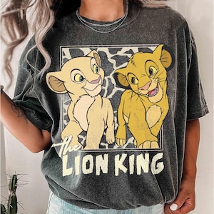 May include: Dark gray t-shirt featuring a graphic of two lion cubs from The Lion King. The cubs are in a rectangular frame with a black and white pattern background. The words "The Lion King" are printed below the cubs.