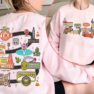 May include: A light pink sweatshirt featuring colorful graphic designs inspired by the movie Cars. The designs include Route 66 signs, cacti, and various shop logos like "Radiator Springs Curios" and "Cozy Cone Motel".
