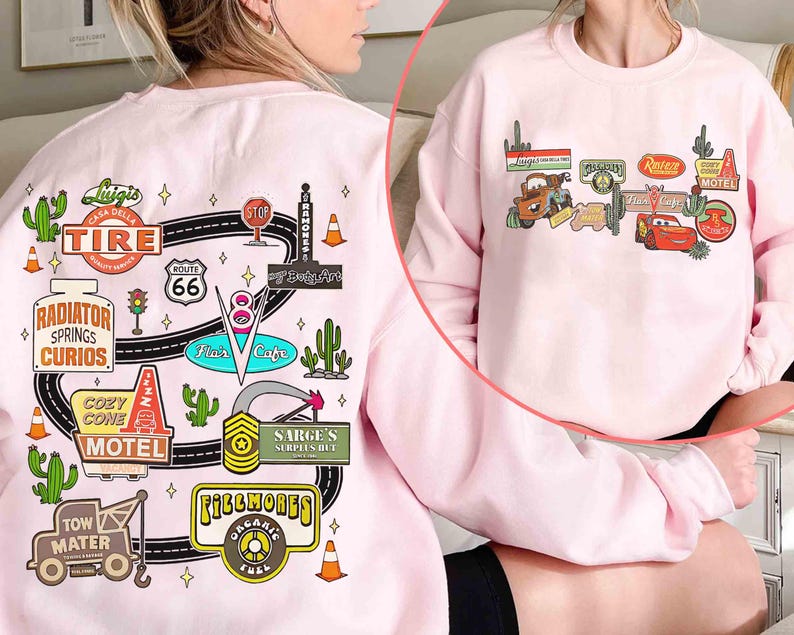 May include: A light pink sweatshirt featuring colorful graphic designs inspired by the movie Cars. The designs include Route 66 signs, cacti, and various shop logos like "Radiator Springs Curios" and "Cozy Cone Motel".