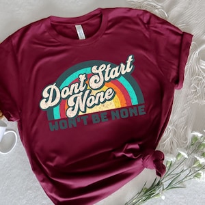 May include: A maroon t-shirt with a vintage rainbow graphic and the text "Don't Start None Won't Be None".