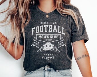 Puede incluir: Una camiseta gris oscuro con un gráfico blanco que dice "Girls Flag Football Mom's Club Est. 2024" con un balón de fútbol y un diseño de laurel. La camiseta también tiene el texto "#6 Robyn" impreso en la parte inferior.