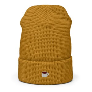 Pode incluir: Um gorro amarelo mostarda com um pequeno design de xícara de café bordado.