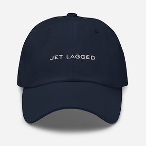 以下が含まれることがあります： 白い刺繍で「JET LAGGED」と書かれたネイビーブルーのベースボールキャップです。